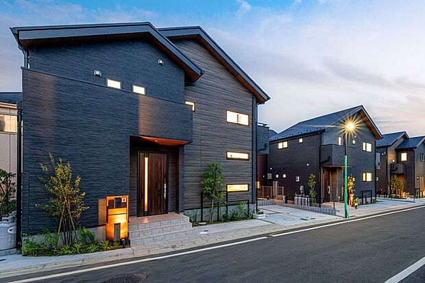 【 住協建設の施工例 】黒の木目調が織りなす重厚な佇まい。 洗練されたダークトーンの質感が夕暮れの街並みに美しく映える、デザイン性の高いモダン邸宅。