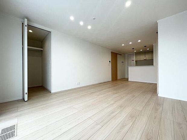 【D棟LDK】明るい木目調のフロアが広がる開放的なLDKです。左奥にはウォークインクローゼット、正面奥にはペンダントライトが灯る対面式キッチンが配置されています。（2025年12月撮影）