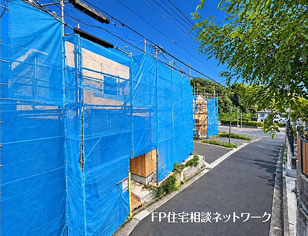 角地に位置する建築中の建物の様子です。周囲は足場と養生シートで覆われ、完成に向けて工事が進められています。接道する道路の状況や隣地との高低差など、周辺の環境が確認できます。