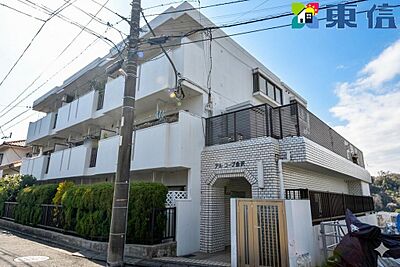 外観：白を基調とした爽やかな外観に、レンガ調のアーチエントランスが映える上品なマンション。手入れされた植栽が優しく包み込み、青空に美しく映える落ち着いた佇まいが日々の暮らしに安心感を添えます。