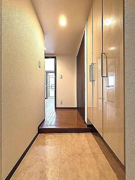天井まで届く大容量シューズボックスを備えた清潔感あふれる玄関空間！トールタイプの収納で家族の靴をスッキリ収められ、落ち着いた木目調とあたたかな照明が住む人やゲストを気持ちよく迎え入れます。