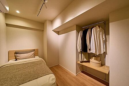 温かみのある木目調のフローリングを採用した洋室には、ハンガーパイプと棚が備え付けられたオープンタイプの収納スペースが設置されており、天井にはダウンライトやスポットライトが配されています。