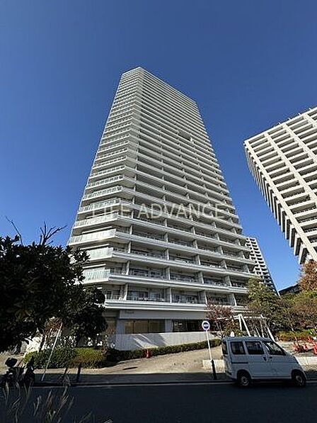 白を基調としたシンプルなデザインの高層マンションで、各階にバルコニーが設置されており、敷地内には植栽や綺麗に舗装されたアプローチ道路が整備されています。