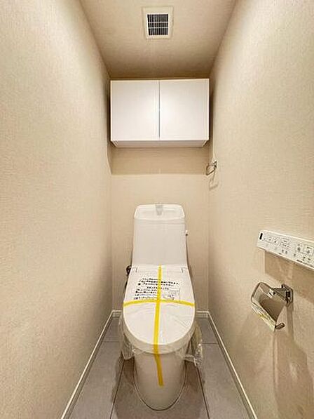 清潔感のあるトイレ空間には、トイレットペーパーなどを収納できる扉付きの吊戸棚が備わっているほか、壁面には操作しやすいリモコンやペーパーホルダー、タオル掛けが設置されています。