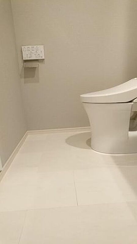 白を基調とした清潔感のあるトイレには、操作しやすい壁付けのリモコンパネルやペーパーホルダーを備えており、落ち着いた色合いのクロスと明るい床面でシンプルに仕上がっています。