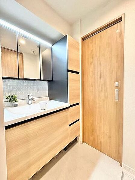 木目調のデザインで統一された洗面室には、三面鏡と上部照明を備えた洗面化粧台に加え、隣接してトールサイズの作り付け収納棚が設置されており、ドアには落ち着いた木目調の引き戸が採用されています。