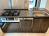 キッチン：石目調のワークトップを採用したシステムキッチンには、3口ガスコンロやビルトインタイプの食器洗い乾燥機を備えており、下部には木目調デザインの収納扉が設置されています。