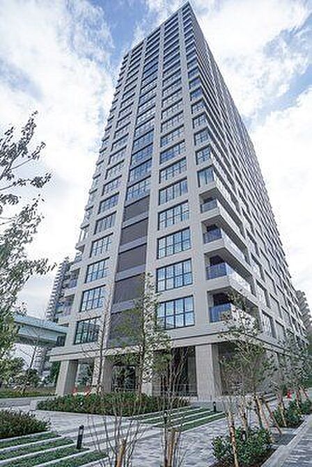 グレーを基調としたモダンな外観デザインの高層マンションで、バルコニーには透明感のあるガラス手すりが採用されており、敷地内には美しく整えられた植栽やタイル敷きのアプローチが整備されています。