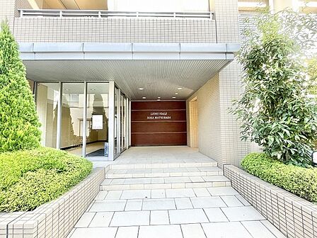 緑の植栽と上品なタイル貼りの外壁が調和する、落ち着きと品格を感じさせるエントランスアプローチ。