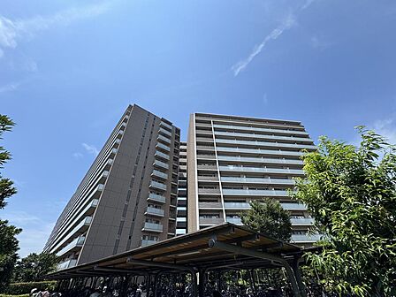 落ち着いた色合いの外壁が青空に映えるマンション外観です。バルコニーが整然と連なる端正なデザインで、敷地内には植栽と屋根付き駐輪場が配置されています。ご家族の帰りを迎える落ち着いたたたずまいです。