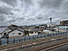 周辺：4階バルコニーからの眺めです。目の前を鉄道の線路が通り、その先には戸建てを中心とした街並みが広がります。前に高い建物がなく、空が広く見えます。※AIによる家具消し加工を行なっております。