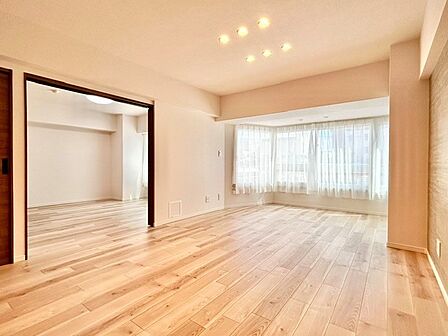 大きな出窓からたっぷりの陽光が届く明るいLDK！引き戸を開放すれば隣接する洋室と一体利用でき、より伸びやかな空間に。美しい木目調フロアとモダンなダウンライトが心地よい暮らしを叶えます。