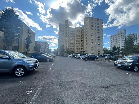 建物の前に広がる平置き駐車場。周囲には木々が植えられ、住まいへ向かう道に緑の彩りを感じられます。車での帰宅や荷物の出し入れを想像しながら、駐車位置や建物までの動線を確認できます。空の広さも印象的です。