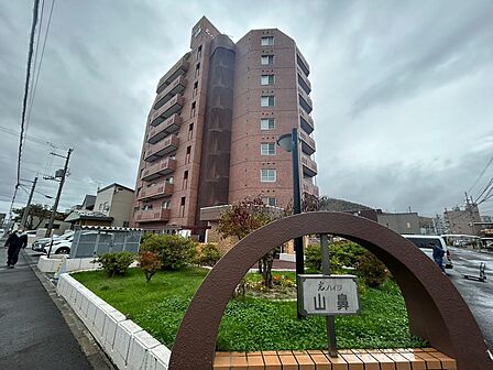 赤茶色の外壁と縦のラインが印象的な光ハイツ山鼻。建物の足元には芝生や植栽が広がり、季節の彩りを身近に感じられます。アーチ型の館名看板も目を引く佇まい。周辺の街並みや建物の出入りも現地でご確認ください。