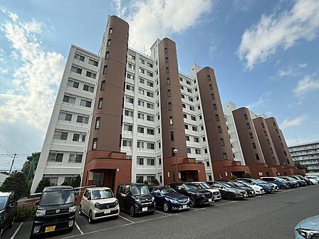 白を基調とした外壁に、ブラウンの縦ラインが映えるマンション外観です。建物前には駐車スペースが広がり、開放感のある佇まいが印象的。青空に映える堂々とした建物の全景から、住まいのスケール感も感じられます。