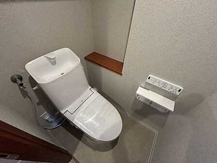 白い便器と落ち着いた壁色が調和するトイレ。温水洗浄便座と壁付けリモコンを備え、快適に使えます。手洗い付きのタンクに加え、背面には小物を置ける棚も。使い勝手と清潔感を大切にした、すっきりとした空間です。