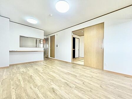 明るい色合いのフローリングが空間全体を広く感じさせます。大型の照明器具が設置されており室内は十分な明るさがあります。開放的な対面式キッチンが隣接し使い勝手の良さが伺えます。