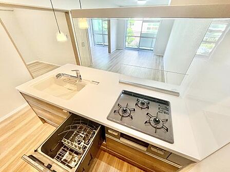 キッチンにはビルトイン食洗機と3口ガスコンロが設置されており調理スペースもゆとりがあります。明るい色合いの床材とカウンターが空間を広く見せます。カウンター越しにリビングが見渡せる設計です。
