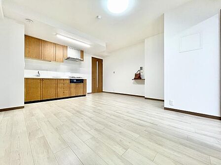 明るいフローリングが印象的なリビングです。キッチンとの一体感があり広々とした空間が広がります。木目調のキッチン収納が落ち着いた雰囲気を演出しています。壁面の白さが清潔感を引き立てます。