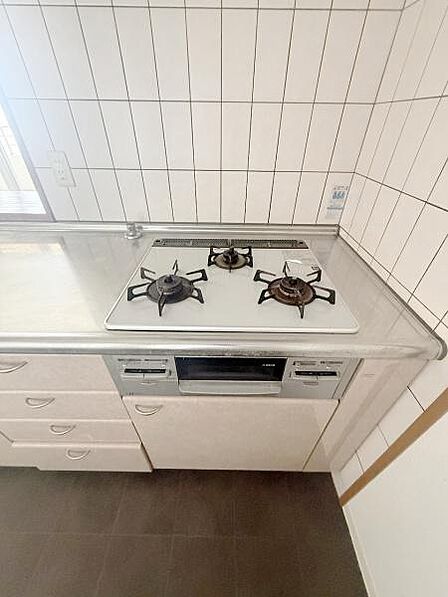 三口コンロが設置されているキッチンです。作業スペースがシンク横に確保されているため調理も進めやすいです。白を基調としたタイル壁で清潔感があります。収納も複数あり整理しやすいレイアウトです。