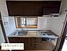 キッチン：シンプルなデザインのキッチンでコンロとシンクが並び作業効率が考えられています。収納スペースもしっかり確保されています。窓から明るい光が差し込み清潔感のあるタイル張りです。