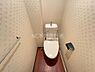 トイレ：温かみのあるフローリング調の床が印象的なトイレです。清潔感のある壁紙とシンプルな設備が魅力です。空間を効率的に活用した設計となっています。ペーパーホルダーやタオル掛けも配置されています。
