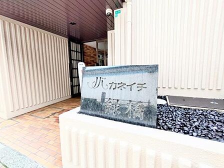 エントランスに設置された石製の建物名表示板が物件の印象を引き締めます。外壁や床は明るい色合いで清潔感のある空間です。エントランスにはセキュリティカメラも設置されています。堅牢な作りが特徴的です。